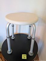 Front view of adjustable bath stool showing white round seat, aluminum legs with height adjustment holes, and rubber feet