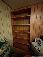 Front view of tall wooden bookcase with five shelves in a room corner next to paneled walls.