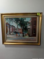 Framed oil painting depicting a streetscape with pedestrians, trees, and buildings, signed by artist Colin Billings, shown hanging on a wall.