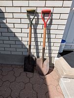Two garden digging tools leaning against a brick wall outdoors on a paved surface, showing front view of handles and blades.