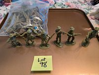 Green plastic army men figures arranged in a line, displayed on a brown surface with a bag of additional figures in the background.