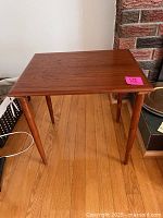 View of the rectangular teak bunching table from an angle showing the wood grain top and tapered legs against a hardwood floor and brick background.