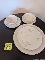 Photo of one place setting with dinner plate, salad plate, dessert plate, small bowl, cup and saucer displayed on black table.