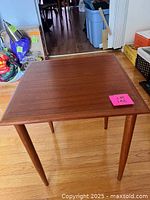 Front angled photo of a small square teak bunching table showing the wooden top and tapered round legs on a hardwood floor.