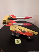 Side profile of the Strenco metal toy rocket launcher transporter with two attached rockets sitting on a black surface, showing yellow vehicle body with red wheels and launcher arm.