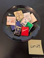 Overhead view of assorted vintage matchbooks placed on a black Jamaican lacquer bowl showing various brand names and logos on matchbook covers.