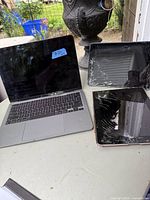 Photo showing 1 MacBook and 2 iPads on a table outdoors. MacBook has screen damage and the iPads have cracked screens.