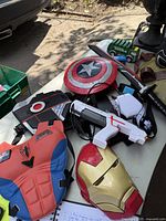 Image of various kids toys and costume pieces on a table including Spider-Man chest armor, Iron Man mask, Captain America shield, and toy guns.