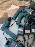 Photo showing 5 teal Makita cordless drills, 4 Makita battery chargers, two cylindrical batteries attached to chargers, and 1 Worx charger with cords on white surface.