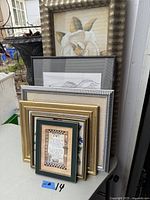 Photo showing six framed wall art pieces stacked against a wall, variety of styles and frame finishes with a botanical print at top