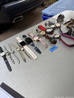 Watches displayed in a row on a white table outdoors near a car and blue recycling bin, showing variety of styles and colors from metal chains to silicone bands.