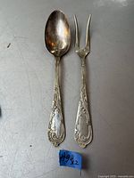 Pair of antique serving fork and spoon with ornate handles and a patina, laid side by side with a small blue paper marked #92