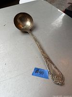 Full view of the antique silver ladle showing the ornate handle and deep bowl sitting on a metal surface with a blue tag labeled #90.