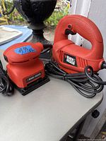 Photo showing Black & Decker palm sander and jigsaw placed side by side on a surface, both red and black in color with power cords.