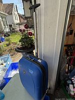 Blue carry-on suitcase with textured exterior, visible telescoping pull-up handle extended, side handle, and wheels at base, photographed in natural light outside next to a white wall and flowers.