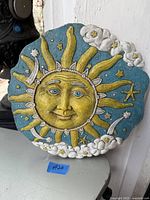 Front view of clay garden wall art depicting sun with facial features, surrounded by sun rays, clouds, stars on blue background.