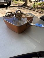 Square copper-colored Copper Chef pan with glass lid on white table outdoors.