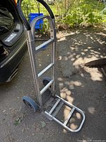 Aluminum hand truck dolly shown from angle with base plate extended, next to a car trunk and garden area.