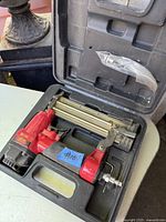 Red and black headless pin nailer placed inside molded black hard plastic case with handle. Clear plastic bag with nails or accessories inside case lid compartment.