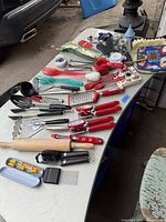 Assorted kitchen tools laid out on a table including red-handled peelers, black serving spoons, graters, can openers, measuring spoons, and other utensils.
