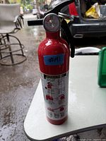 Red fire extinguisher with black handle and pressure gauge sitting on white surface outdoors. Label and blue sticker with #M1 visible.