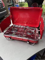 Open double burner camp stove showing two burners, metal grate, and red case with lid and side wind guards.