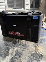 Front and side view of black Skip The Dishes delivery bag with handles visible.