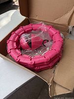 Pink segmented plastic weighted hula hoop coiled in box with attached pink and black weighted ball wrapped in plastic.