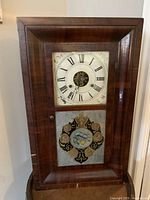 Full front view of antique wooden mantel clock showing rectangular shape, decorative painted clock face with Roman numerals, and lower door with ornate painted glass panel.