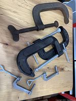 Top-down view of five different metal clamps on a wood surface including large vintage C-clamp and smaller black and silver clamps.