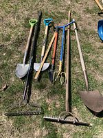 Overview of seven assorted outdoor garden tools laying on grass showing different types and sizes of shovels, rakes, and garden tools.