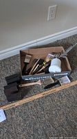 Box contains assorted paint brushes, sanding blocks, and a handheld paint sprayer placed on carpet next to ruler for scale.