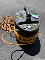 Top and front view of yellow and black Bostitch air compressor with pressure gauges and control knob visible, orange attached hose coiled beside compressor.