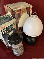 Black propane lantern with white frosted glass globe next to its box and a Coleman battery-operated lantern in original box