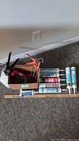 Box containing two caulking guns and multiple tubes of caulk on floor against wall outlet