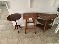 Three occasional tables arranged side by side on tiled floor under white shelves: round pedestal table, square table, half-moon two-shelf table