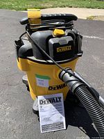 Full view of yellow and black DeWalt wet/dry vacuum standing on asphalt with attached hose and instruction manual propped against the base.