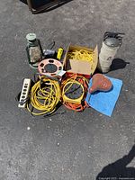 Full lot overview showing all items on concrete ground including extension cords, bike seat, lantern, flashlight/radio, oil reservoir, blue tarp, hand brush.