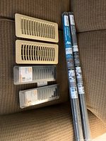 Seven floor registers and screening rolls displayed on a brown chair upholstery, showing two black HDX floor registers still in plastic packaging labeled 3 x 10 inches, two beige used floor registers, and two rolled screen packages.