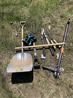 Photo showing a selection of garden tools including a large shovel, garden fork, hedge clippers, hedge trimmer, weed puller, sprinkler, and wooden measuring stick arranged on grass.