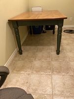 Side view of rectangular harvest table showing natural finish wooden tabletop and turned forest green leg in room with tiled floor.