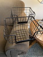 Two black metal wire baskets placed on a cushioned chair armrest and wooden table, showing handles and open wire design.