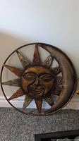 Metal sun and moon wall decor in rust color, measuring about 20x20 inches, showing detailed facial features and crescent moon with several points, resting on carpeted floor.