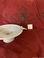 Photo of small white tabletop bird bath with bird figure on rim and one metal decorative wall hanging lying on red fabric.