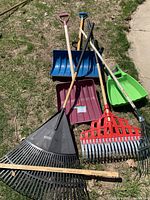 Front view of assorted outdoor equipment including snow shovels and leaf rakes arranged on grass and sidewalk.