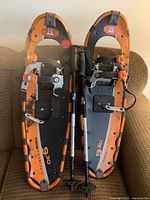 Top view of pair of orange and black Yukon Charlie’s 930 snowshoes with adjustable bindings and two telescopic walking poles placed between them on a brown couch.