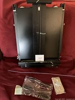 Photo of the portable black easel folded flat, showing a rectangular piece with knobs on sides for adjusting, set against a red background.