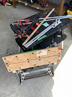 Photo of two portable work tables stacked with assorted pipe clamps and a Mastercraft Acu Rip tool on top. The wooden Black & Decker workbench is folded and bottom, the black plastic portable table on top.