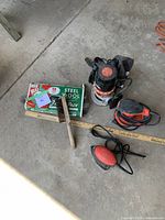 Top-down view of power tools on concrete floor including round and triangle Black & Decker sanders, Sears router, steel wool box, and brush.