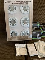 Photo showing unopened package of Commercial Electric 3-inch recessed lighting 6-pack containing six can lights with white trims. Also visible are instruction sheets and mounting hardware.